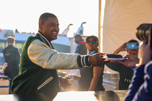 A man smiling in a green varsity jacket while giving away a bowl of food.