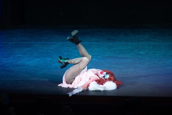 A drag queen with long red hair poses on her back in the middle of a stage.