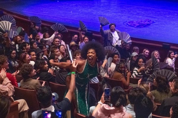 A drag queen in a long green dress stands in the middle of the audience while many audience members hold open fans up.