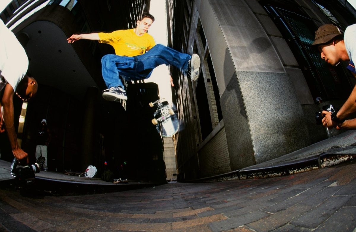 A man in a yellow shirt with blue jeans does a skateboarding trick over the camera. Two men are filming him.