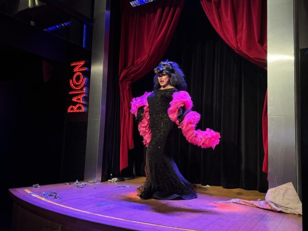 A drag queen stands on stage with a hot pink feather boa, a black sequin dress, and a hat shaped like a wolf head.