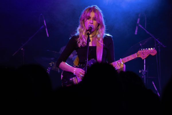 A woman singing into a microphone and playing a guitar, looking straight into the camera.