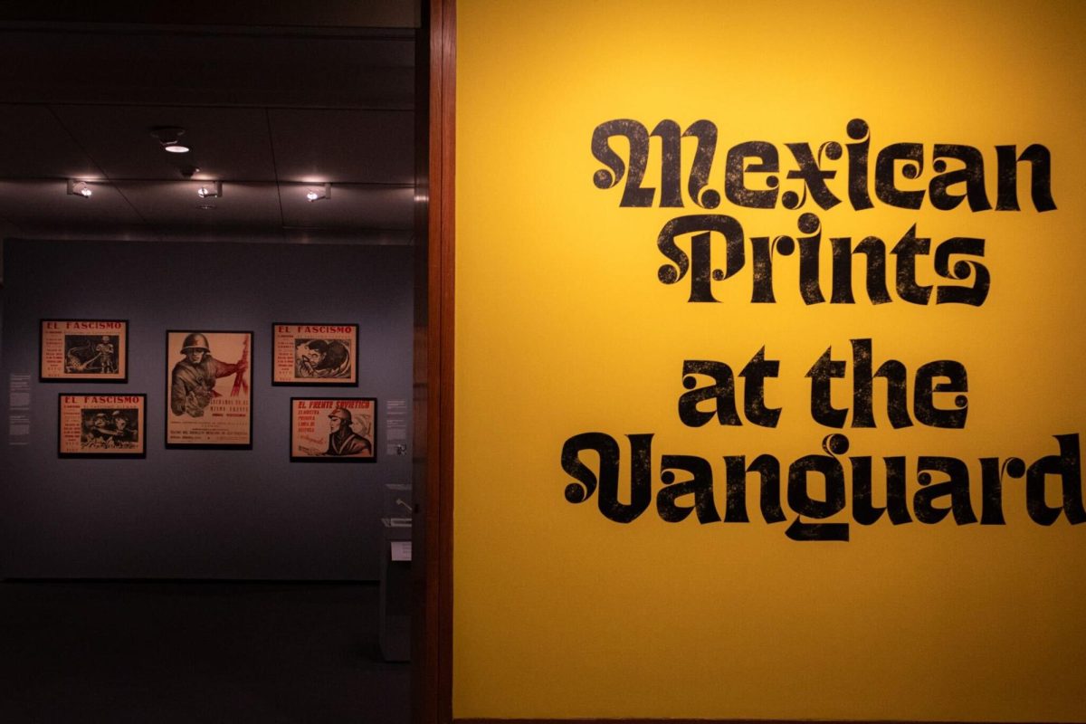 An exhibit featuring a bold yellow wall that says “Mexican Prints at the Vanguard” with the posters displayed on the wall in the background.