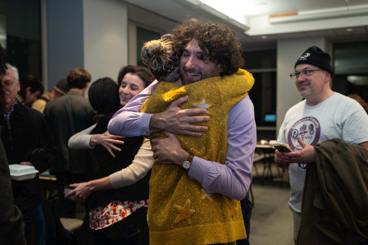 Two people hugging in a crowded room, while others in the background also hug and smile.