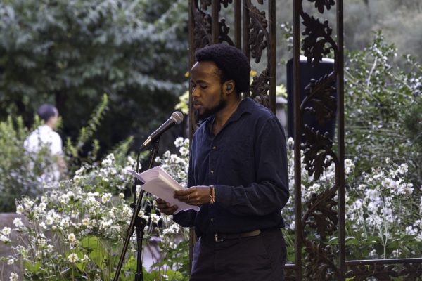 A man is speaking at a microphone, holding a piece of paper, surrounded by flowers and bushes.