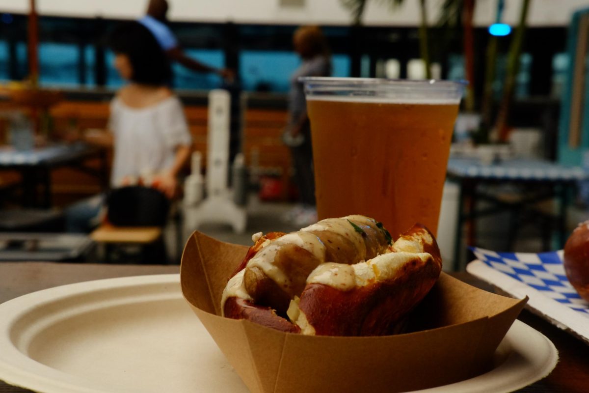 A close-up shot of a bratwurst with white sauce on top in a cardboard tray, in front of some beer.