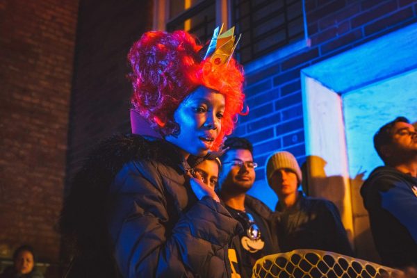 A woman in costume wears a pink wig with a crown. She has her hand over her chest, and is illuminated by blue light.