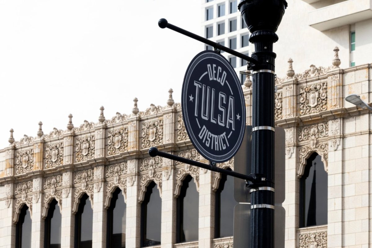 A black lamppost sign that reads “TULSA” and “DECO DISTRICT” with beige, stone buildings behind it.