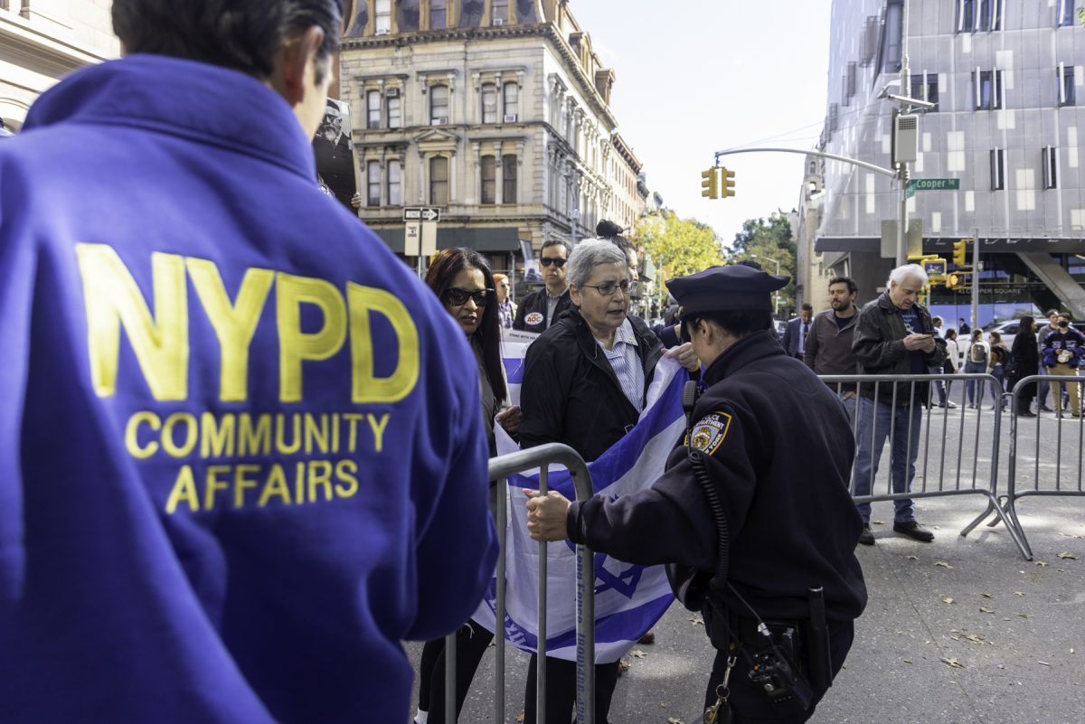 A police officer holds onto a metal barricade, blocking protesters holding Israeli flags from a space. A person in the foreground watches and the back of his jacket reads: “N.Y.P.D. COMMUNITY AFFAIRS.”