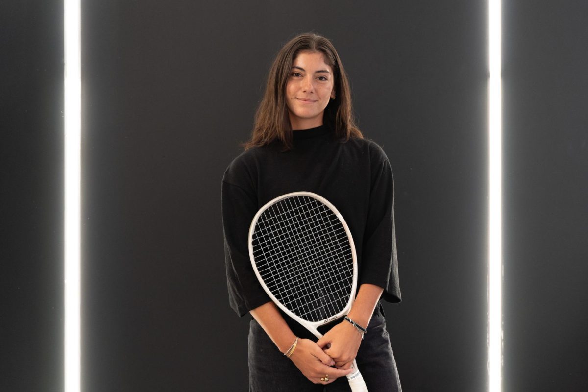 A person stands against a dark gray background with two L.E.D. light strips on either side and is wearing all black as she holds a tennis racquet.