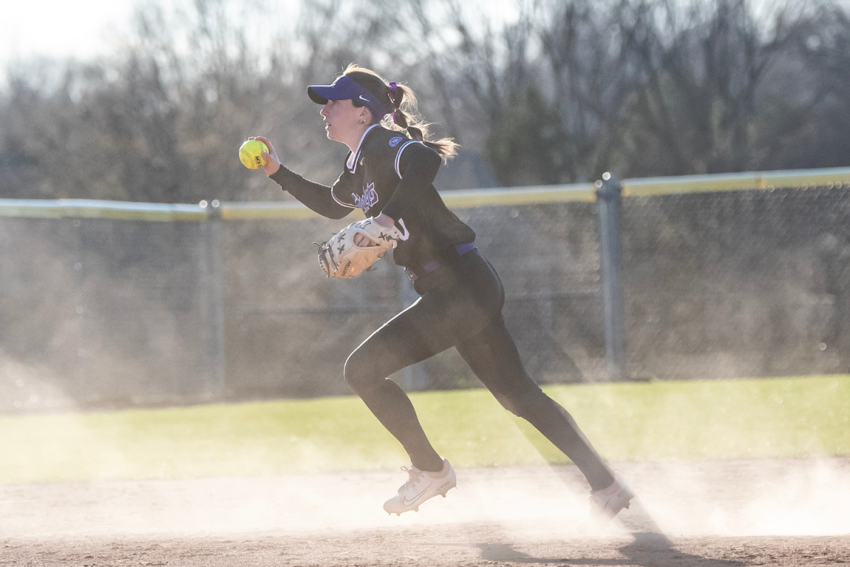 A girl running with a yellow ball in her hand and a softball glove on her other hand. There is a lot of dust at her feet.