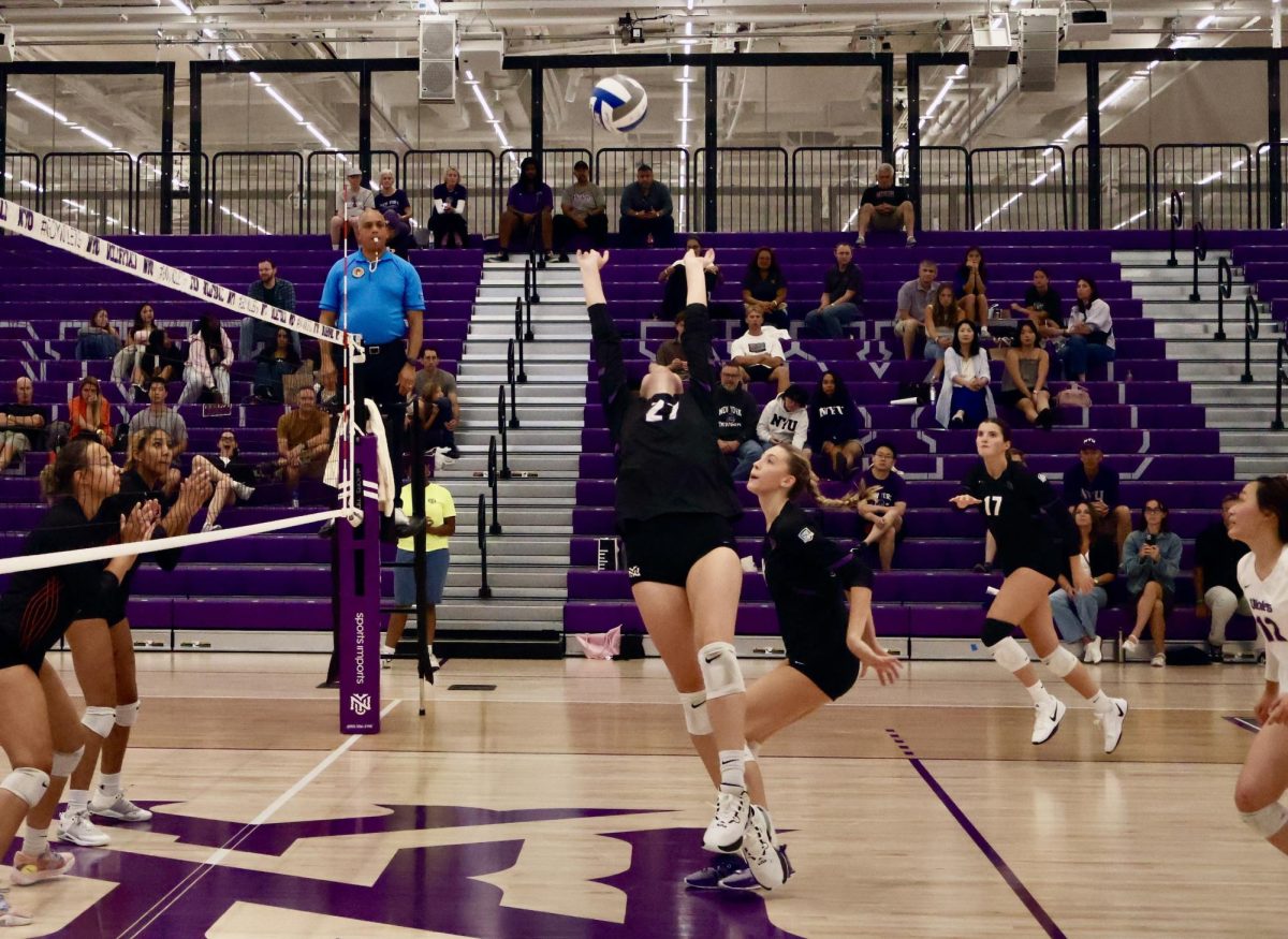 Three volleyball players stand on one side of the net looking at a ball in the air, while a fourth player jumps in the air to set the ball.