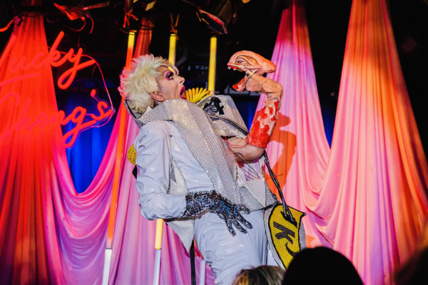 A drag queen stands back in shock while wearing a white suit and a sequin glove on one hand. On the other hand, an alien puppet pops out from the middle of the suit.