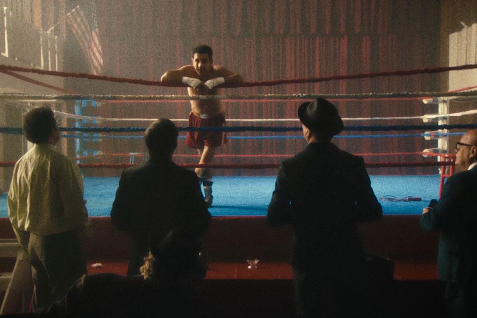 Four people look up to a fighter who is in a boxing ring. The fighter is hanging on the ropes, looking back down to the others.