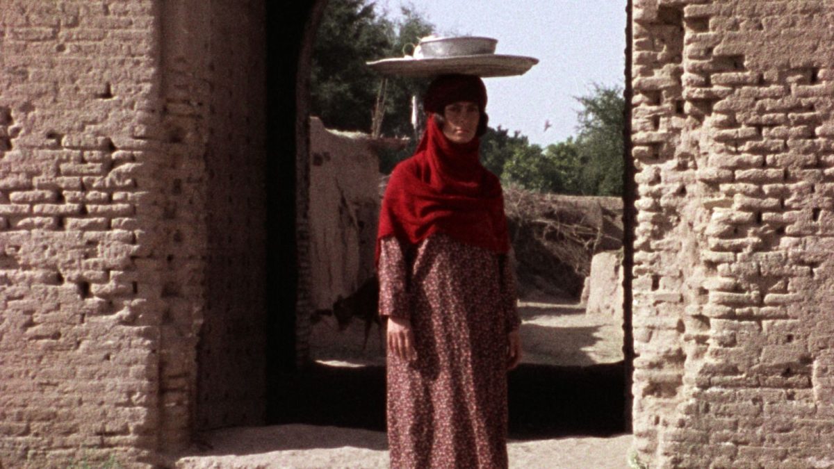 A woman wearing a red headscarf with a long brown dress, while balancing a tray of bowls and cups in her head, standing around an old brick gate.