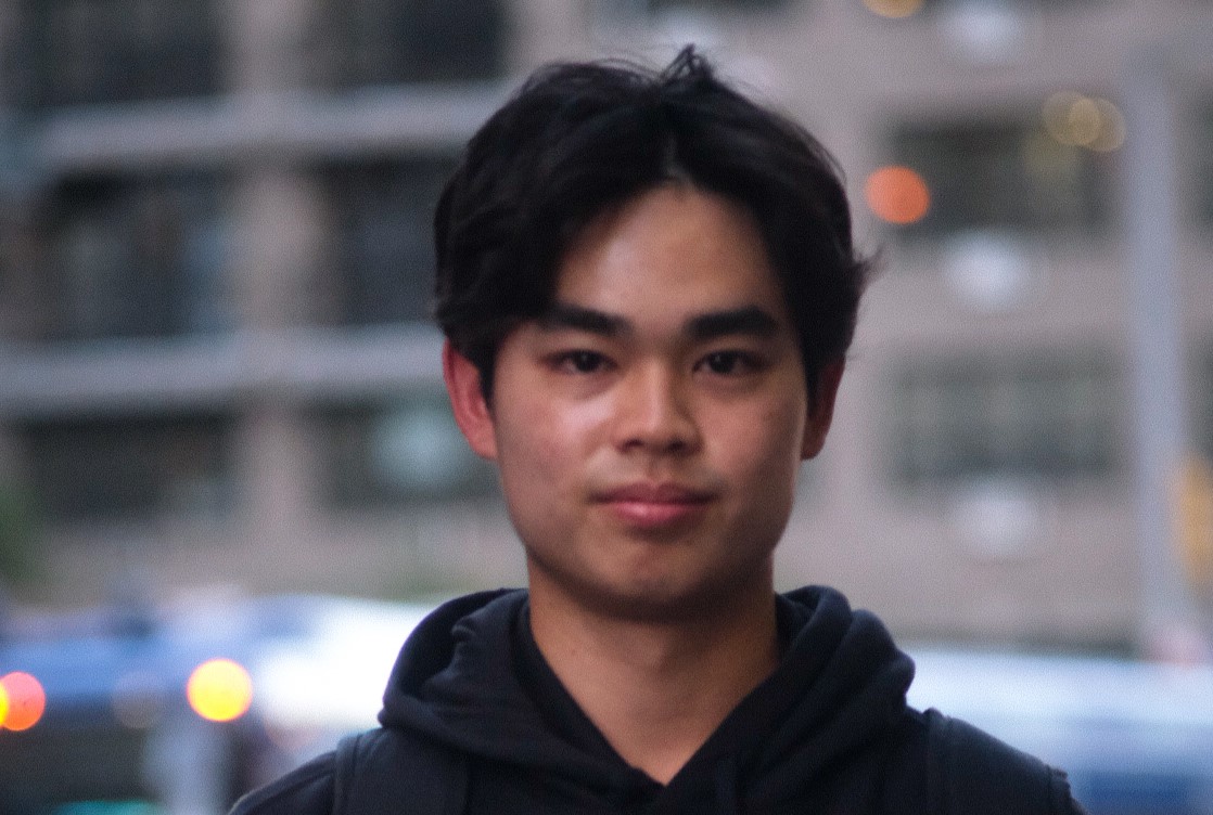 A dark-haired athlete in a black hoodie faces the camera standing on a city street.