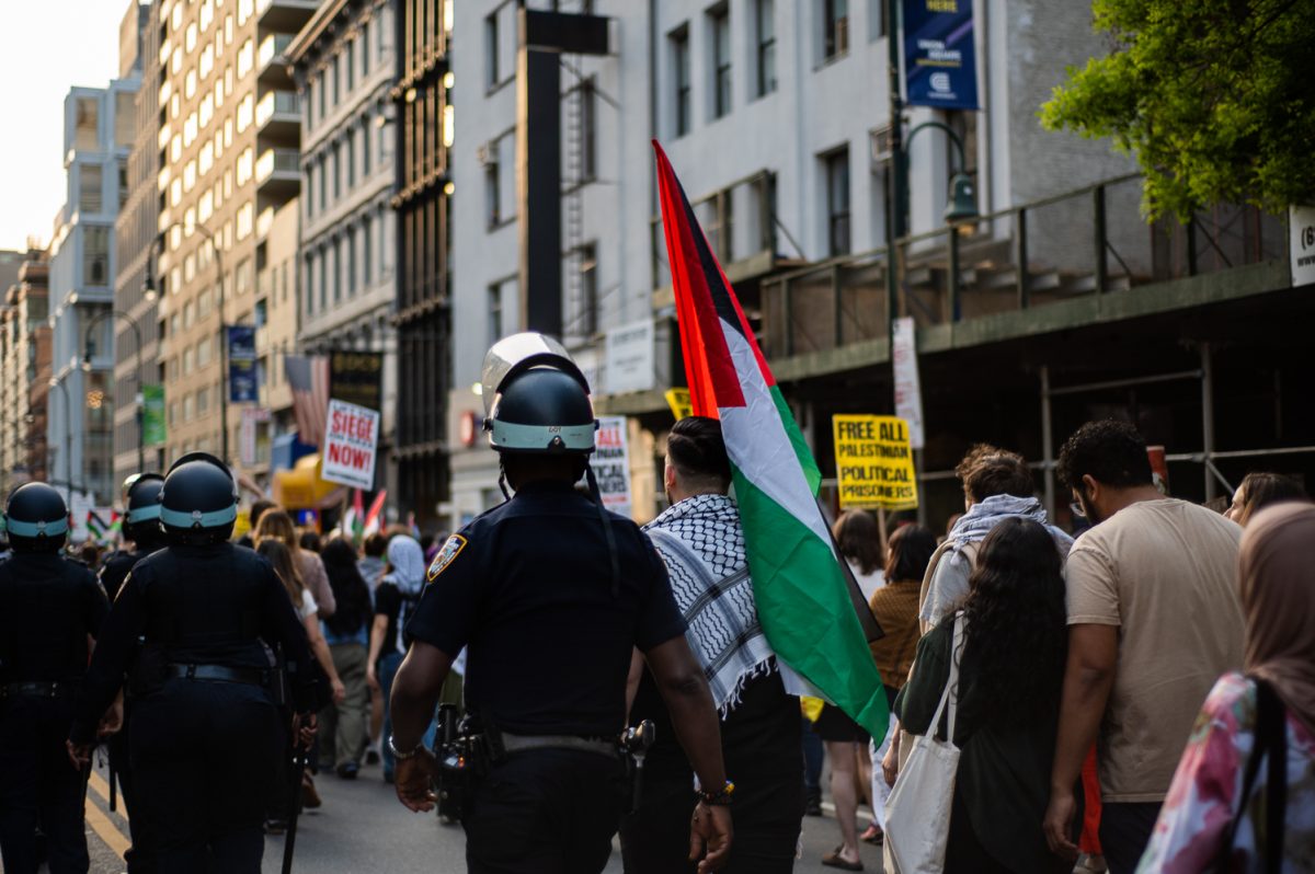 A group of protestors walk down the street, the person in the middle holding a Palestinian flag. On the left side there are police officers walking.