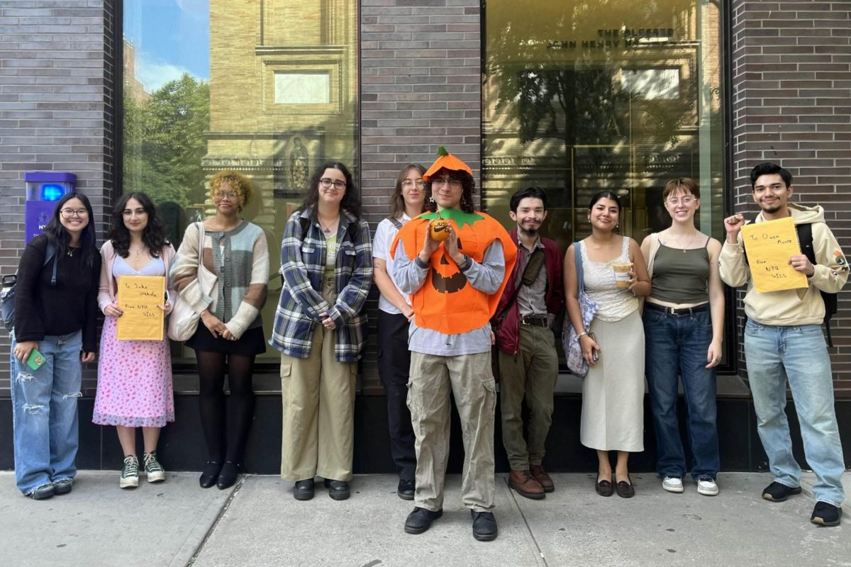 N.Y.U. students stand in a row. Two students hold yellow envelopes. A student in the middle wears a pumpkin costume.