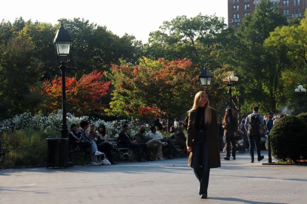 A woman walking alone in the park, looking to her left. There are people sitting on the bench and walking in the background.