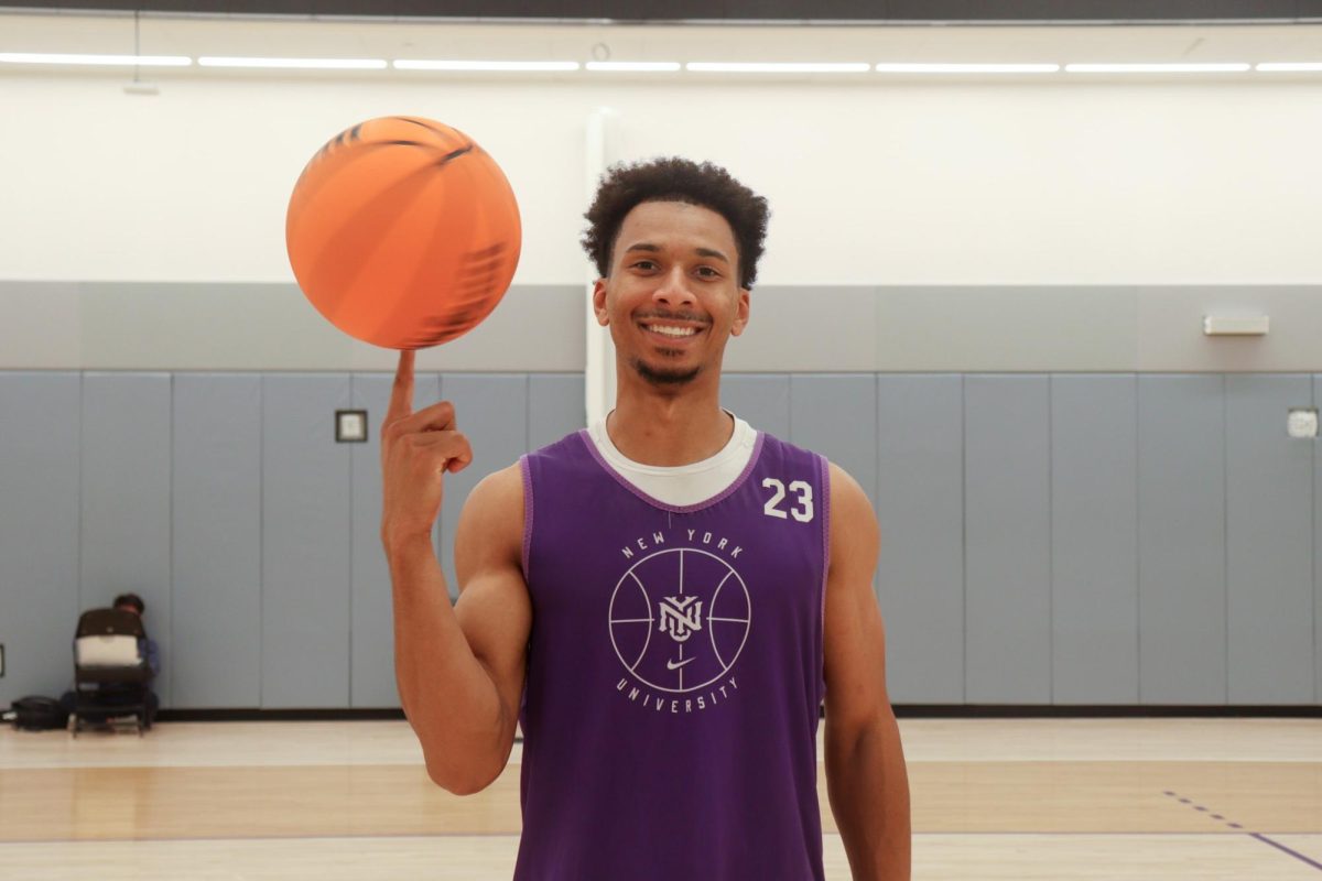 A man wearing a purple “New York University” jersey has a spinning basketball on his fingertip, smiling at the camera.