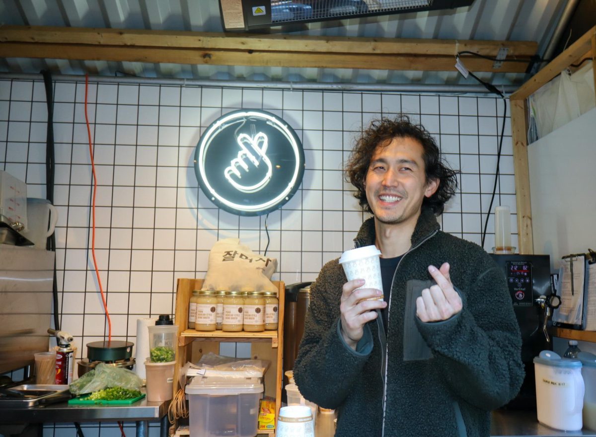 A man stands inside a food stall, smiling. Behind him is a neon sign with a logo of a finger heart.