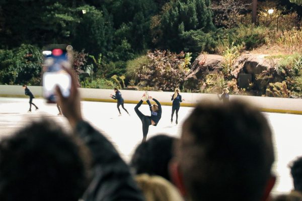 A person in a crowd films a group of figure skaters performing on an ice rink.