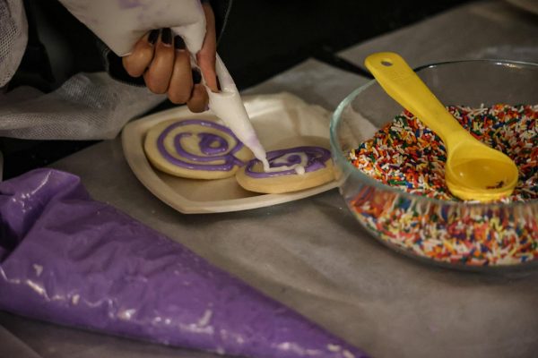 Two cookies on a white plate with purple and white frosting in a swirl. A bowl of sprinkles sits beside the plate.