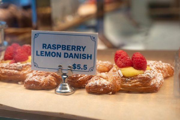 A pastry topped with powdered sugar, yellow custard and raspberries. There is a small sign that reads “Raspberry Lemon Danish $5.5” in front of the pastry.