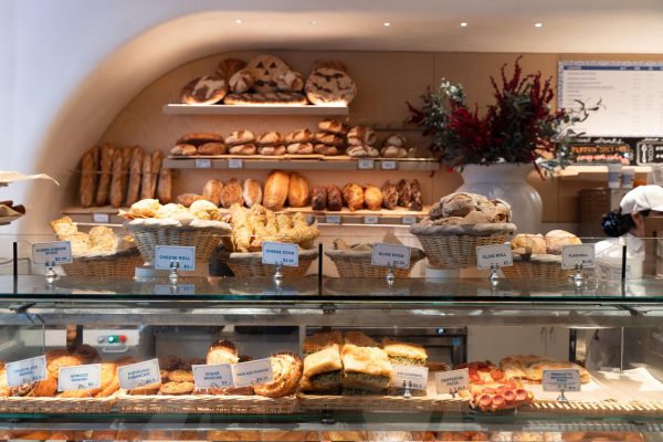 : Baskets of assorted pastries behind a glass, with shelves of bread loaves on the back wall.