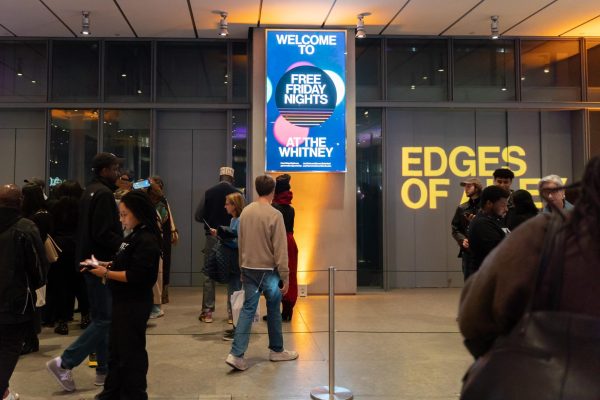 A white room contains an electronic screen in the middle that says “WELCOME TO FREE FRIDAY NIGHTS AT THE WHITNEY” while a crowd waits in line.