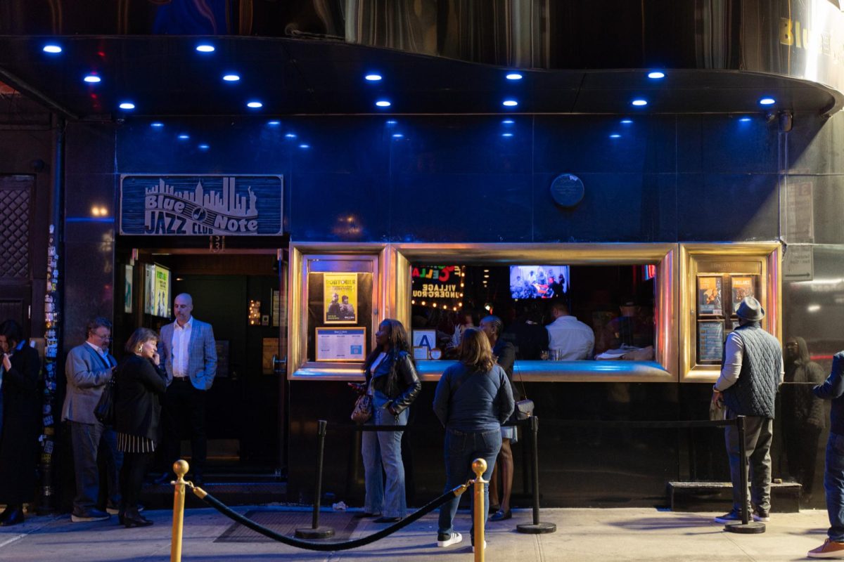 The exterior of a building with blue lighting and an awning shaped like a piano. On the left side is a rectangular wooden sign that says “Blue Note Jazz Club,” with gold windows on the right