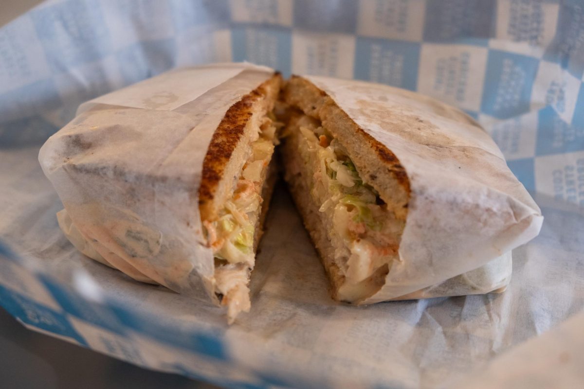 A sandwich wrapped in paper, filled with turkey, cheese and coleslaw, sitting in a blue-and-white checkered paper wrap.