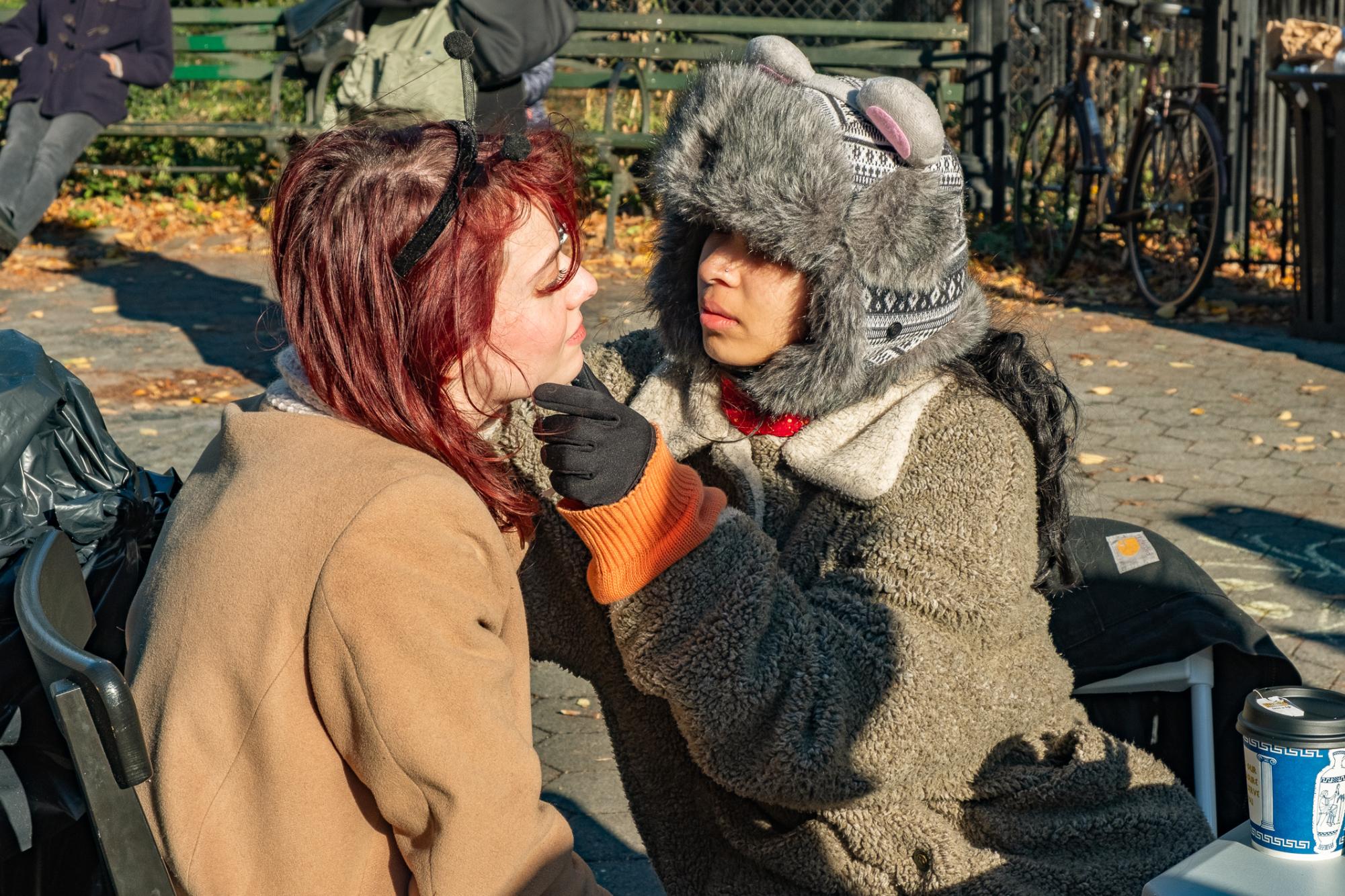 A person in a furry hood and mouse ears paints someone else’s face in a park.