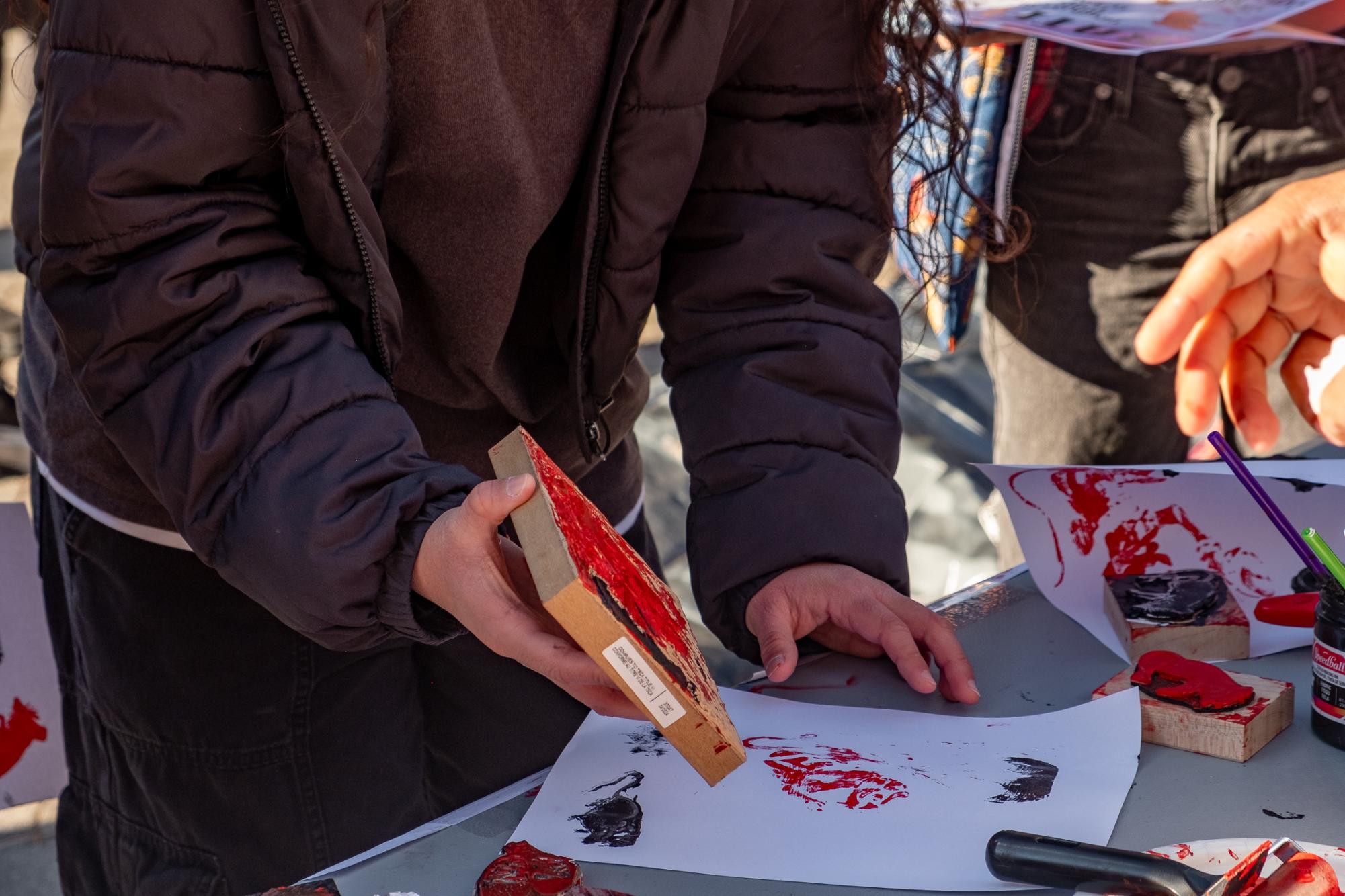 A person stands over a table holding a wooden block with red paint on it. On the table is a sheet of paper with a red design in paint.