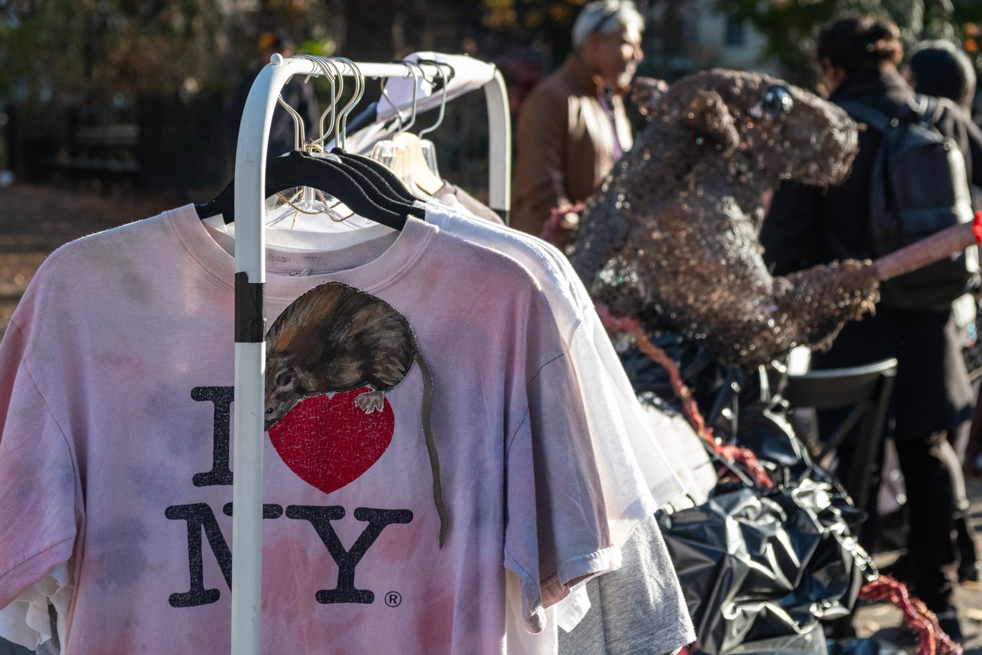 A rack of shirts in a park. The one in front says “I heart N.Y.” with a painted rat on it.