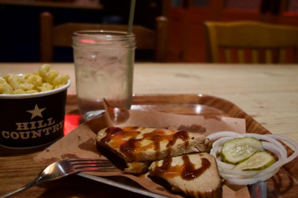 A serving of smoked turkey breast with barbecue sauce on a plate, accompanied by pickles, onions and a mac ‘n’ cheese side, with a mason jar of water on a wooden table.