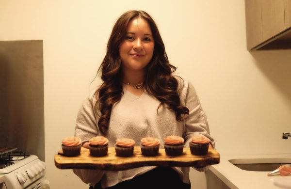 A woman with wavy brown hair holds a tray of pink-frosted chocolate cupcakes.