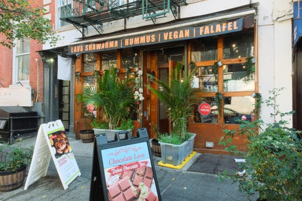 A wooden exterior of a restaurant with plants in the front. A black awning above the entrance says “LAVA SHAWARMA, HUMUS, VEGAN, FALAFEL” in orange block letters.