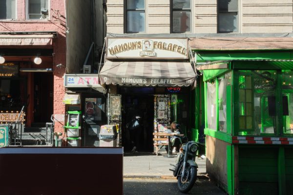 The exterior of a restaurant from across the street with a striped awning with the name “MAMOUN’S FALAFEL.”  The name also appears on a white and brown sign above the awning.