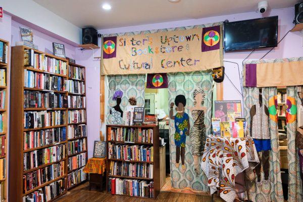 The interior view of a shop with wooden shelves filled with books. A colorful fabric banner hangs overhead that says “Sister’s Uptown Bookstore and Cultural Center.” Below are patterned curtains with African-inspired figures.