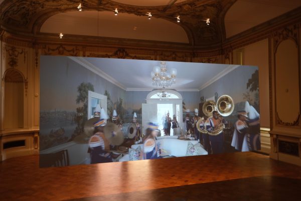 A hanging screen in an empty room, playing a video of a marching band performing around a dining room table.