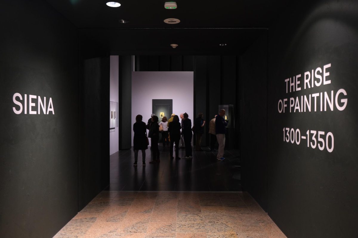 A hallway that leads into an exhibit is painted black and dimly lit. The wall to the left says “SIENA” and the wall to the right says “THE RISE OF PAINTING, 1300-1350.”