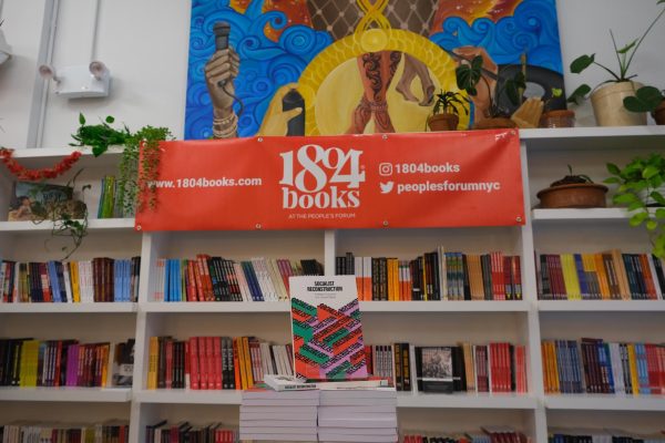 The interior view of a store contains a white bookshelf filled with colorful books. A red banner hanging above shelves reads “1804 books.” A vibrant mural of a microphone and cultural elements is on the wall behind the shelf, and a stack of books titled "Socialist Reconstruction" is displayed in front.