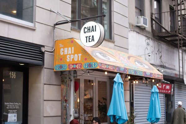 An exterior of a Chai Tea shop with yellow awning and colorful artwork with the sign reading "The Chai Spot", located under a building. 