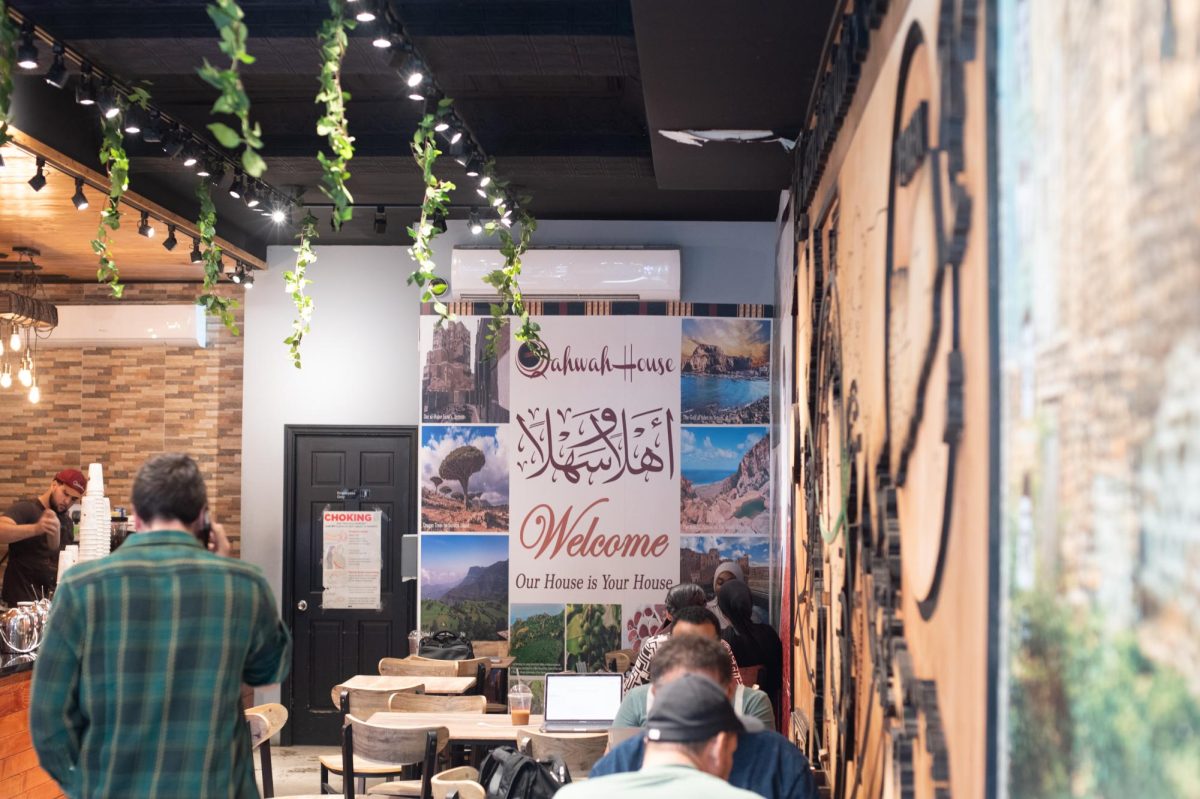 A look into the interior of a restaurant with people eating inside. In the back, a poster says “Qahwah House Welcome, Our House is Your House” next to several images of nature.