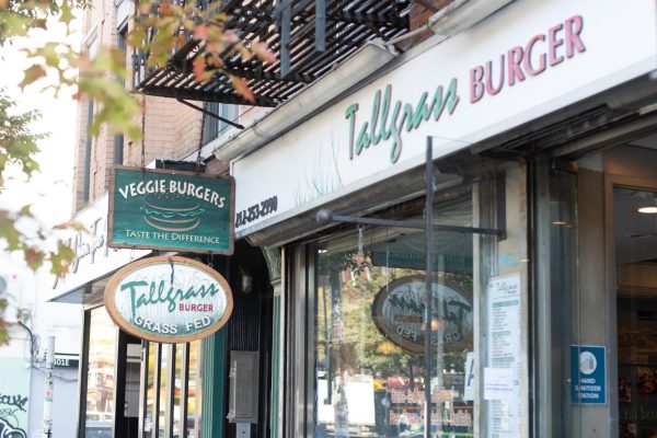 The front of a restaurant with the words "Tallgrass Burger" above the door. Hanging off the restaurant is a sign with a burger that reads “VEGGIE BURGERS TASTE THE DIFFERENCE” and “Tallgrass BURGER GRASS FED.”