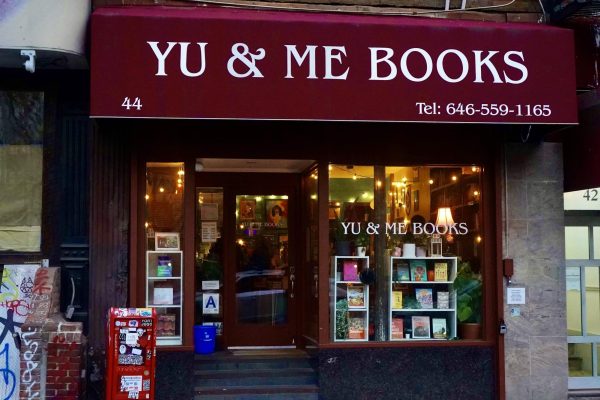 An exterior view of a bookstore with red awning that says “YU & ME BOOKS” and the store's phone number. The entrance is made up of wooden door frames and large windows showcasing books and warm lighting.