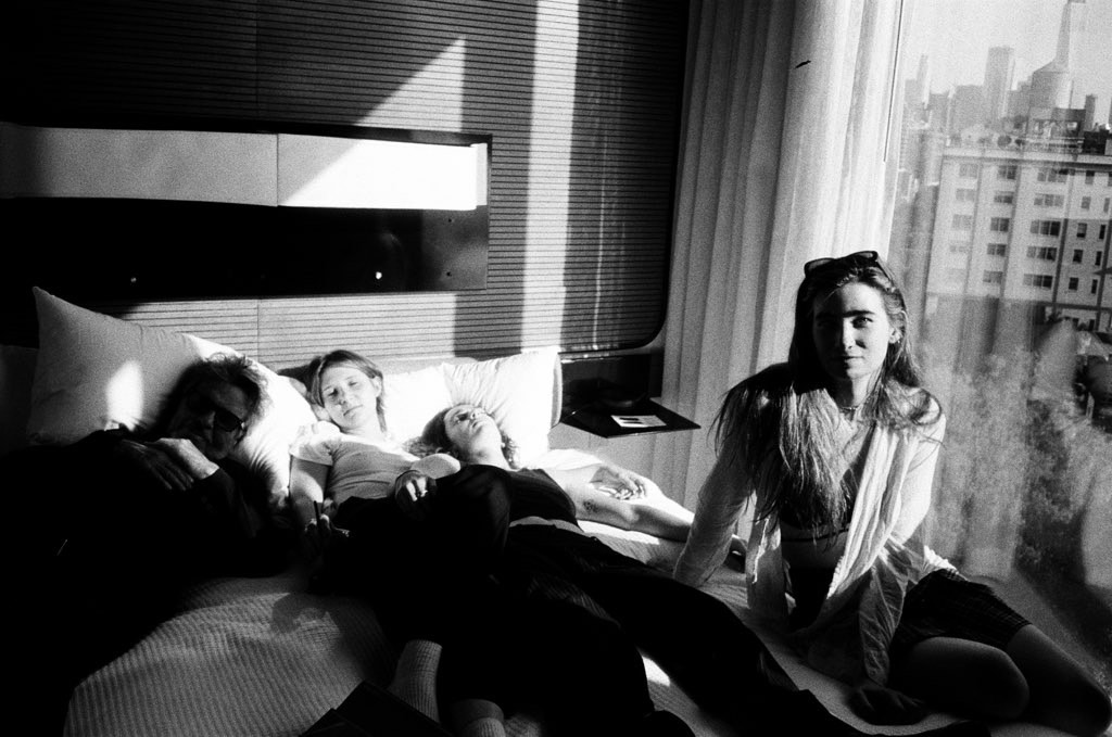 A black-and-white picture of three people lying on a bed, one person sits up, looking at the camera.