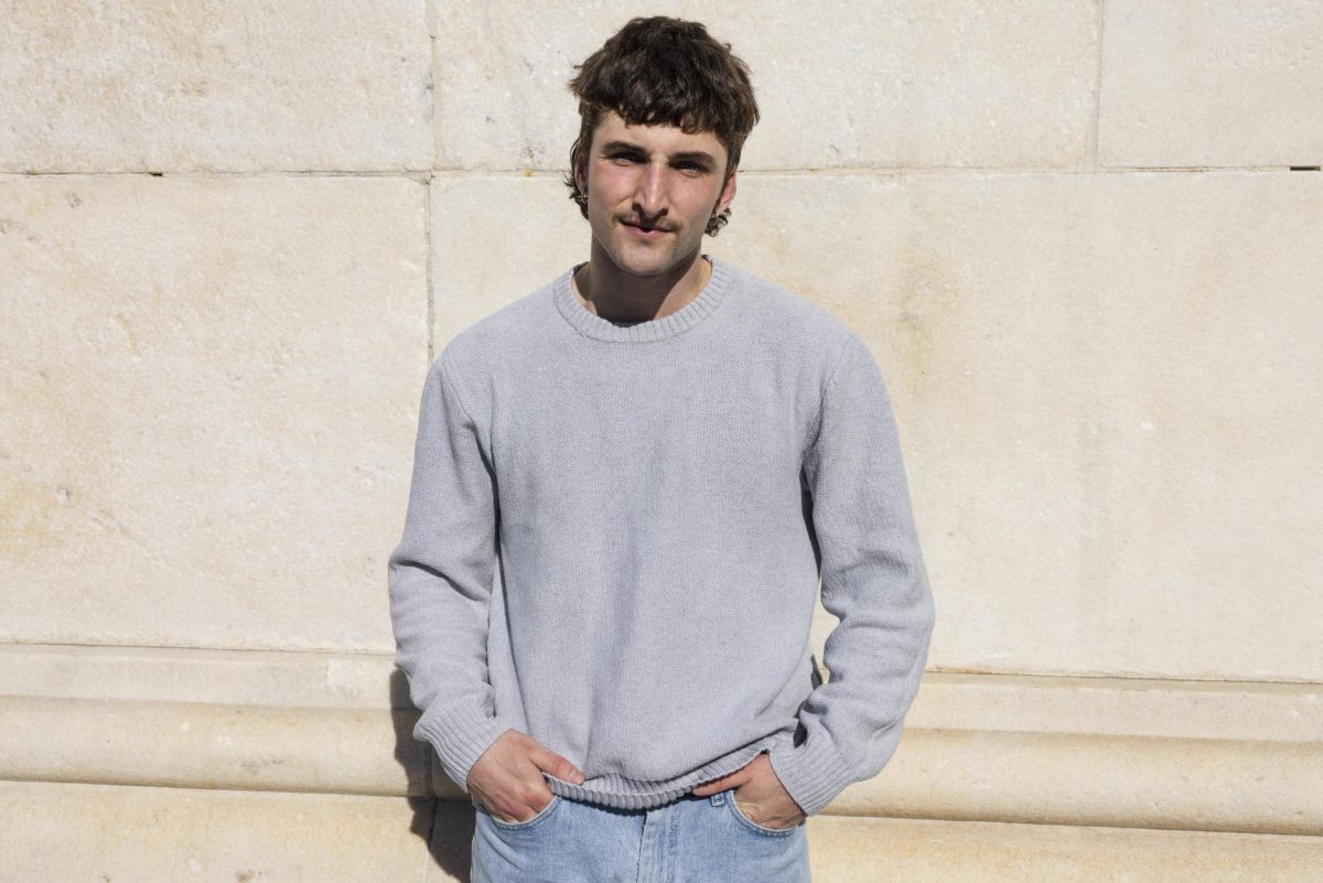 A man in a gray crewneck and jeans puts his hands in his pockets while looking at the camera.