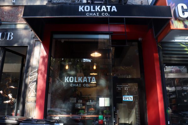 An exterior of a shop with black awning and red walls. In the middle is a glass window that has a sign that says “KOLKATA CHAI CO.”